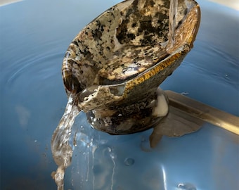 May include: A decorative stone bowl with water pouring into it. The bowl has a speckled pattern of brown, black, and white. The water flows out of the bowl into a blue surface.