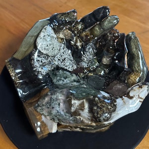 May include: A hand-shaped bowl made of ceramic and decorated with embedded stones. The bowl is a light brown color with green, black, and white stones embedded in the surface. The bowl is sitting on a black surface.