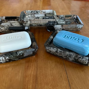 May include: Two soap dishes made of black and white speckled resin with a raised rim. One dish has a white bar of soap that says "Dove" and the other dish has a blue bar of soap that says "Coast".