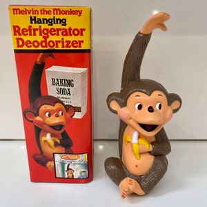 Peut inclure: Une figurine de singe marron tenant une banane jaune, suspendue à un crochet. Le singe est un désodorisant pour réfrigérateur, comme l'indique la boîte en arrière-plan, qui indique "Melvin the Monkey Hanging Refrigerator Deodorizer".