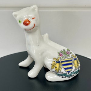 May include: White ceramic figurine of a cat with a winking green eye and a red nose. The cat is sitting with its legs tucked under its body. The figurine has a blue and yellow crest on its back with the text "Civitas Nove Sarum Salisbury".
