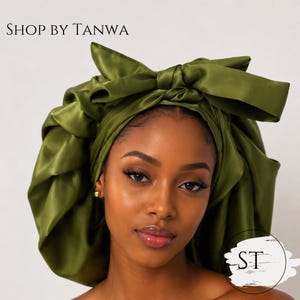 May include: An olive green satin hair bonnet featuring a large decorative bow tied at the top. The text SHOP BY TANWA appears in the upper left corner, and a circular logo with the letters ST is visible in the lower right corner.
