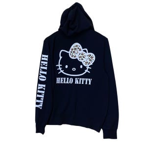 May include: Black hooded sweatshirt featuring a white Hello Kitty graphic on the back with a leopard print bow. The words "HELLO KITTY" are printed below the graphic. The left sleeve has the words "HELLO KITTY" printed vertically in white.