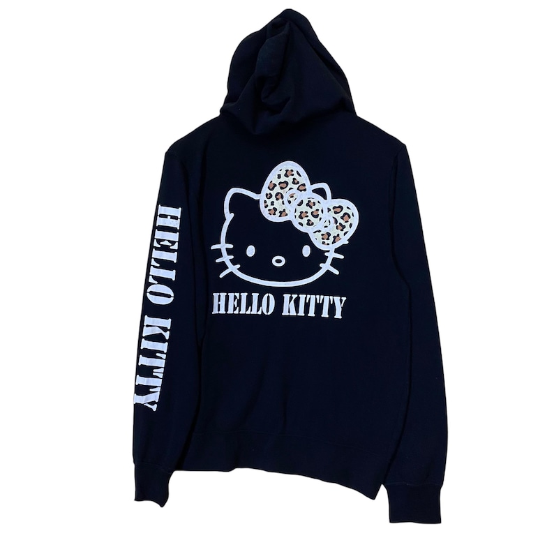 May include: Black hooded sweatshirt featuring a white Hello Kitty graphic on the back with a leopard print bow. The words "HELLO KITTY" are printed below the graphic. The left sleeve has the words "HELLO KITTY" printed vertically in white.