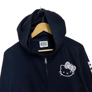 May include: Black zip-up hoodie featuring a Hello Kitty graphic with a leopard print bow. The hoodie has a drawstring hood, a full-length zipper, and the Hello Kitty logo on the chest. The size tag reads "M".