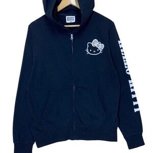 May include: Black zip-up hoodie featuring a white Hello Kitty graphic on the chest and the brand name on the hood. The right sleeve displays the Hello Kitty name in white. The hoodie has a front zipper and two front pockets.