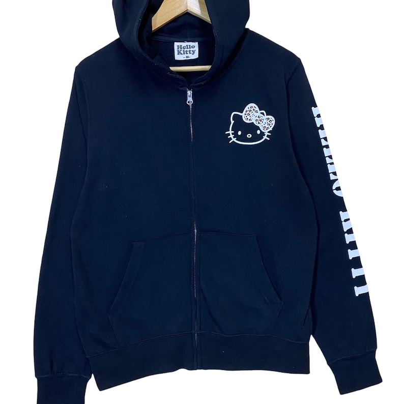 May include: Black zip-up hoodie featuring a white Hello Kitty graphic on the chest and the brand name on the hood. The right sleeve displays the Hello Kitty name in white. The hoodie has a front zipper and two front pockets.