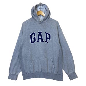 Könnte beinhalten: Grauer Kapuzenpullover mit dem Wort "GAP" in marineblauen Großbuchstaben auf der Brust. Der Pullover hat eine Kängurutasche und eine Kapuze mit Kordelzug. Das Kleidungsstück hängt an einem Kleiderbügel vor weißem Hintergrund.