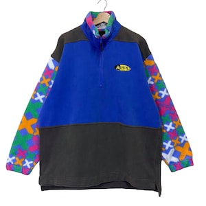 May include: A color-blocked fleece pullover with a blue body, gray bottom, and patterned sleeves. The sleeves feature a colorful cross pattern. The pullover has a quarter-zip closure and the word "APPI" in a yellow oval.