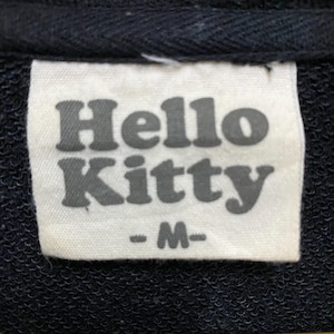 May include: Close-up of a white tag with the words "Hello Kitty" in gray, block letters. Below the text is the letter "M" in gray, indicating the size. The tag is sewn onto a black fabric.