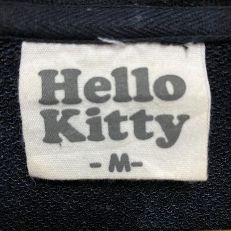 May include: Close-up of a white tag with the words "Hello Kitty" in gray, block letters. Below the text is the letter "M" in gray, indicating the size. The tag is sewn onto a black fabric.