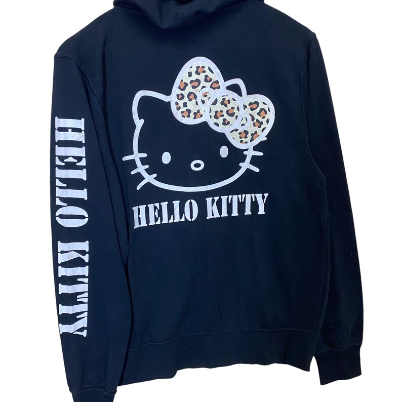 May include: Black hooded sweatshirt featuring a white Hello Kitty graphic with a leopard print bow. The words "HELLO KITTY" are printed in white on the back and down the left sleeve. The sweatshirt has a drawstring hood.