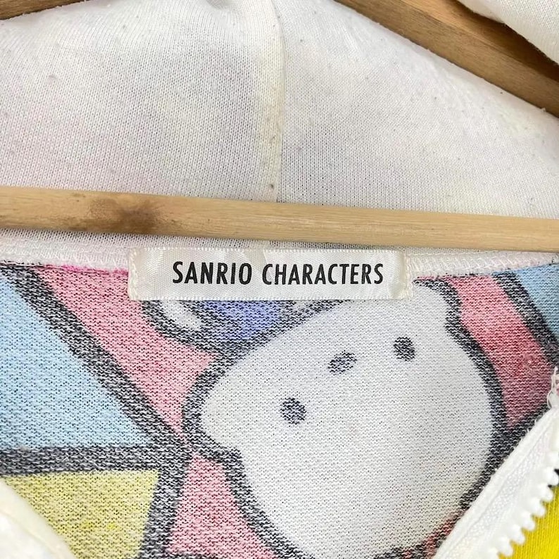 May include: A white hooded sweatshirt with a colorful pattern and the text "SANRIO CHARACTERS" on a tag. The design includes pink, blue, and yellow elements. The zipper is yellow.