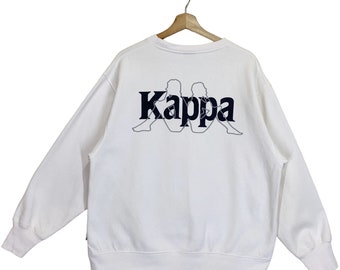 Könnte beinhalten: Weißer Pullover mit einem dunkelblauen Kappa-Logo auf der Rückseite. Das Logo zeigt die charakteristische Silhouette der Marke Kappa von zwei Personen, die Rücken an Rücken sitzen.