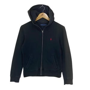 May include: A black zip-up hoodie featuring a hood, long sleeves, and two front pockets. The garment has a small red embroidered logo on the left chest and a blue interior neck label with the text Ralph Lauren printed in white lettering.