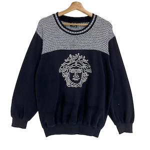 Puede incluir: Una sudadera azul marino con un estampado de cuadros blancos y negros en los hombros y un diseño de cabeza de Medusa blanca en la parte delantera. La sudadera tiene mangas largas y cuello redondo.