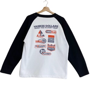 Könnte beinhalten: Weißes und schwarzes Raglan-Ärmel-Shirt mit dem Text "YANKEE DOLLARS ROCK THE PLANET TOUR" und verschiedenen Logo-Designs. Das Shirt hängt an einem Holzbügel. Die Ärmel sind schwarz, der Körper ist weiß.