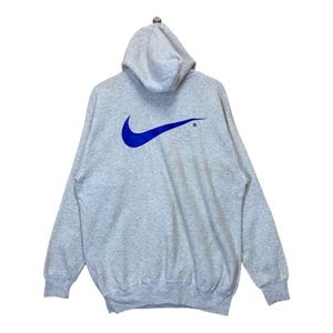 May include: A light grey hoodie featuring a large blue Nike swoosh logo on the back. The hoodie has a drawstring hood and long sleeves. The Nike logo is a prominent design.