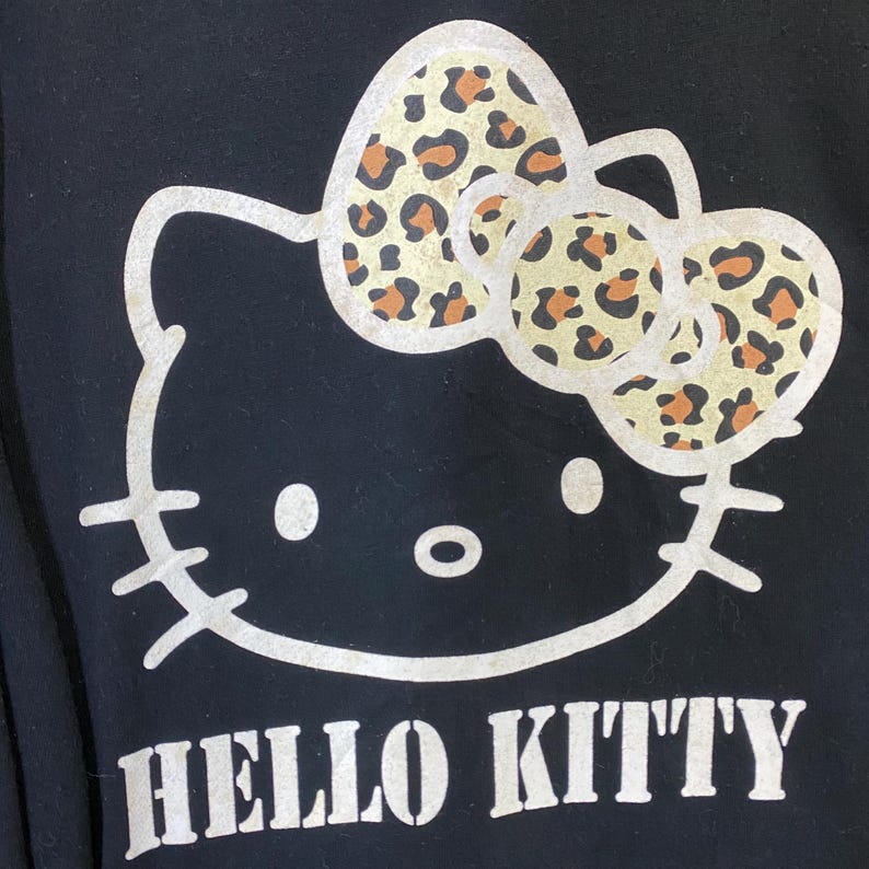 May include: Black garment featuring a white Hello Kitty graphic. The iconic character's bow has a tan and brown leopard print pattern. The words "HELLO KITTY" are printed in white block letters below the character.
