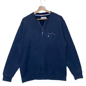 Puede incluir: Un suéter azul oscuro de manga larga con un solo botón en el cuello y un pequeño bolsillo en el pecho izquierdo. El suéter tiene una textura acanalada.