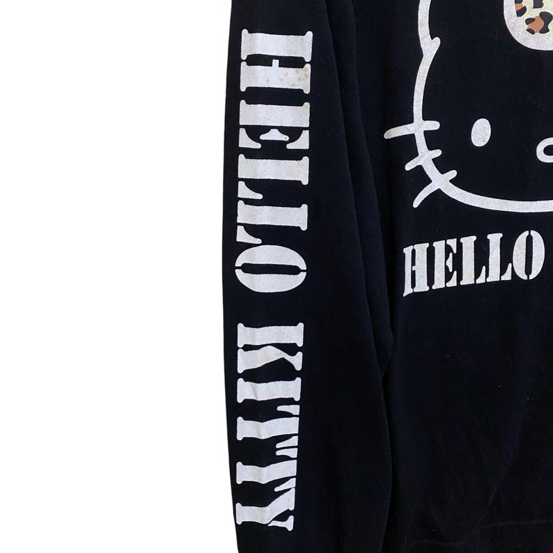 May include: Black sweatshirt featuring a white Hello Kitty graphic and the word "HELLO" in white. The left sleeve has the words "HELLO KITTY" printed vertically in white. The sweatshirt has a leopard print bow.