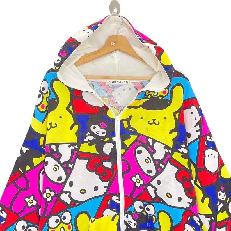 May include: A colorful zip-up hoodie featuring a vibrant pattern of cartoon characters in yellow, pink, blue, and white. The hoodie has a white hood lining and a wooden hanger. The text "SANRIO CHARACTERS" is visible on the label.