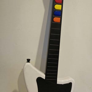 May include: A white and black plastic video game guitar controller featuring a long neck with five colored fret buttons in light blue, red, yellow, dark blue, and orange. The body includes a black pickguard, a strum bar, and two circular buttons.