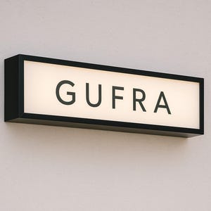 May include: A rectangular illuminated sign with the word "GUFRA" in a bold, sans-serif font. The sign has a black frame and is mounted on a white wall. The light is a soft, warm tone.