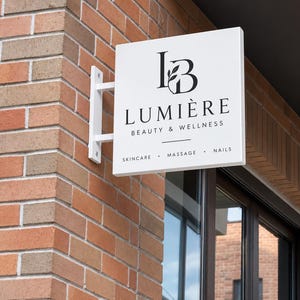 May include: A square white projecting sign mounted on a brick wall. The sign features a black logo with the letters LB and a leaf, the text LUMIÈRE BEAUTY & WELLNESS, and the words SKINCARE, MASSAGE, and NAILS separated by dots in black lettering.
