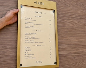 May include: A rectangular menu board with a gold-toned metal frame and a cream-colored insert. The text reads ALBINI, MENU, STARTERS, BRUSCHETTA, BURRATA SALAD, GRILLED OCTOPUS, MAINS, PASTA AL POMODORO, CHICKEN MILANESE, GRILLED SALMON, DRINKS, and more.