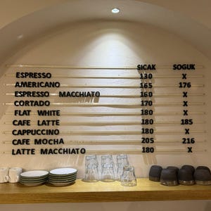May include: A cafe menu board with various coffee drinks listed, including Espresso, Americano, and Latte Macchiato. The menu also lists prices for hot and cold beverages. Below the menu are glasses, cups, and saucers on a wooden shelf.
