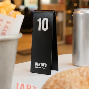 May include: A restaurant table setting featuring a black table number 10, a silver can, a burger, and a container of fries. The table number has the text "Barto's Burger Place". The burger has a sesame seed bun.