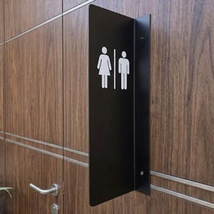 May include: A black metal wall-mounted restroom sign featuring white pictograms of a person in a dress and a person in trousers, separated by a vertical line. The sign is mounted on a dark wood-paneled wall with horizontal silver trim accents.