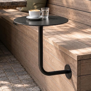 May include: A matte black circular metal side table mounted directly to a wooden bench. The table features a single curved support arm attached to the bench slats and holds a white ceramic coffee cup on a saucer and a clear glass of water.