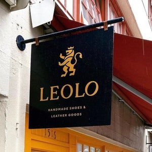 Puede incluir: Un letrero rectangular negro con letras doradas y un logotipo de león dorado. El letrero dice "LEOLO HANDMADE SHOES & LEATHER GOODS". La tienda tiene una puerta y marcos de ventanas amarillos, con un toldo rojo encima.