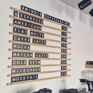 May include: A wall-mounted menu board featuring horizontal wooden rails holding black rectangular tiles with white text. The menu lists items including Espresso, Americano, Flat White, Caffe Latte, Mocha, Matcha Latte, Cold Brew, Milkshake, and Mocktail.