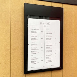 Black outdoor restaurant menu box mounted on a wood paneled wall. Modern wall menu display cabinet with a transparent protective cover for cafe menus, breakfast selections and commercial food price lists.