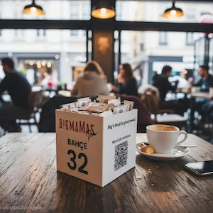 Puede incluir: Escena de café con una mesa de madera. Sobre la mesa, una caja blanca con "BIG!MAMAS BAHÇE 32", una taza de café, un platillo y un smartphone. El fondo muestra personas borrosas.