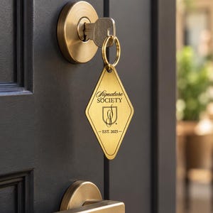 Könnte beinhalten: Ein rautenförmiger Schlüsselanhänger aus Messing, der an einem silbernen Schlüssel in einem Messingschloss hängt. Der Anhänger zeigt den eingravierten Text Signature SOCIETY, ein Schild-Logo mit den Buchstaben SS und EST. 2025.