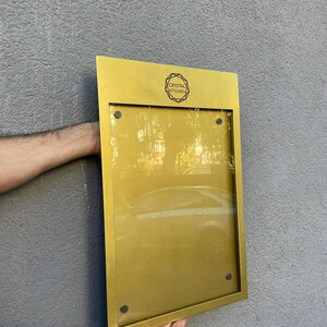 Gold metal wall menu display case with a customized upper section and transparent acrylic front. Personalized restaurant menu cabinet for premium dining venues, hotel restaurants and hospitality entrance signage.