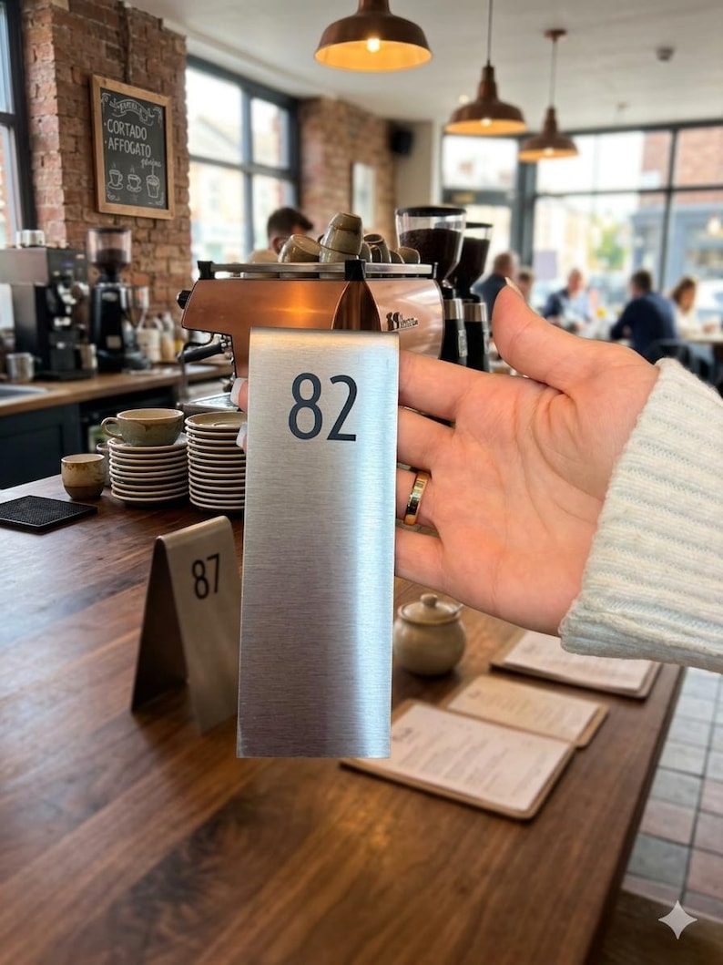 May include: A brushed metal table number with the number 82 in black. The number is held in front of a cafe setting with a wooden table, espresso machine, and other cafe items.