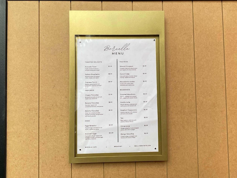 Gold metal menu display frame with a white printed restaurant menu and neatly arranged food categories. Decorative wall mounted menu holder for upscale restaurants, boutique hotels, bistros and dining room interiors.