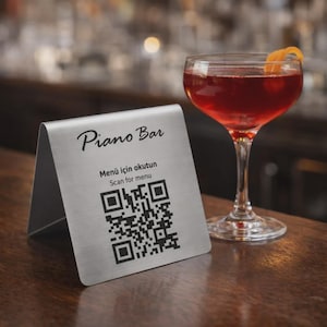 May include: A brushed metal table tent with the words "Piano Bar" and a QR code for a menu. A cocktail with an orange peel garnish sits next to the tent on a wooden bar.