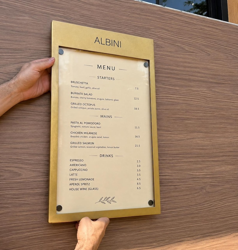 Gold wall mounted restaurant menu display case with a printed food and drinks menu. Elegant metal menu holder with a clear acrylic front, designed for restaurant entrances, hotel dining areas and bistro menu presentation.