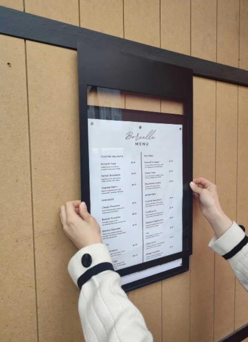 Black metal menu display frame with a transparent front panel protecting a printed restaurant menu. Wall mounted price list holder suitable for cafes, food counters, bars and commercial dining spaces.