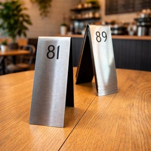 以下が含まれることがあります： 木製のテーブルの上に、番号81と89が表示された2つのつや消し金属製のテーブル番号サインがあります。数字は太字の黒いフォントです。背景には、柔らかな照明のあるカフェの様子が写っています。