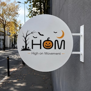 Peut inclure: Une enseigne murale circulaire blanche présentant une silhouette noire d'arbre dénudé et de pierres tombales, deux chauves-souris noires, un croissant de lune orange, le texte HOM avec une citrouille d'Halloween et High on Movement.