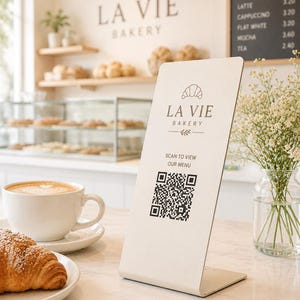 Pode incluir: Uma placa de acrílico cor de creme em forma de L com o logótipo La Vie Bakery, um ícone de croissant e um código QR com o texto "SCAN TO VIEW OUR MENU". A placa está colocada sobre uma superfície de mármore junto a uma chávena de café.