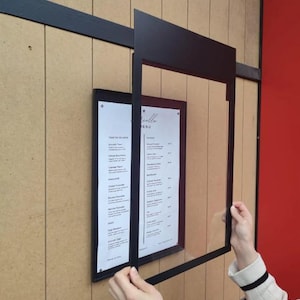 Black wall mounted menu frame with a removable clear acrylic front panel being positioned over a printed menu. Practical restaurant menu display case designed for updating food selections, seasonal menus and price lists.