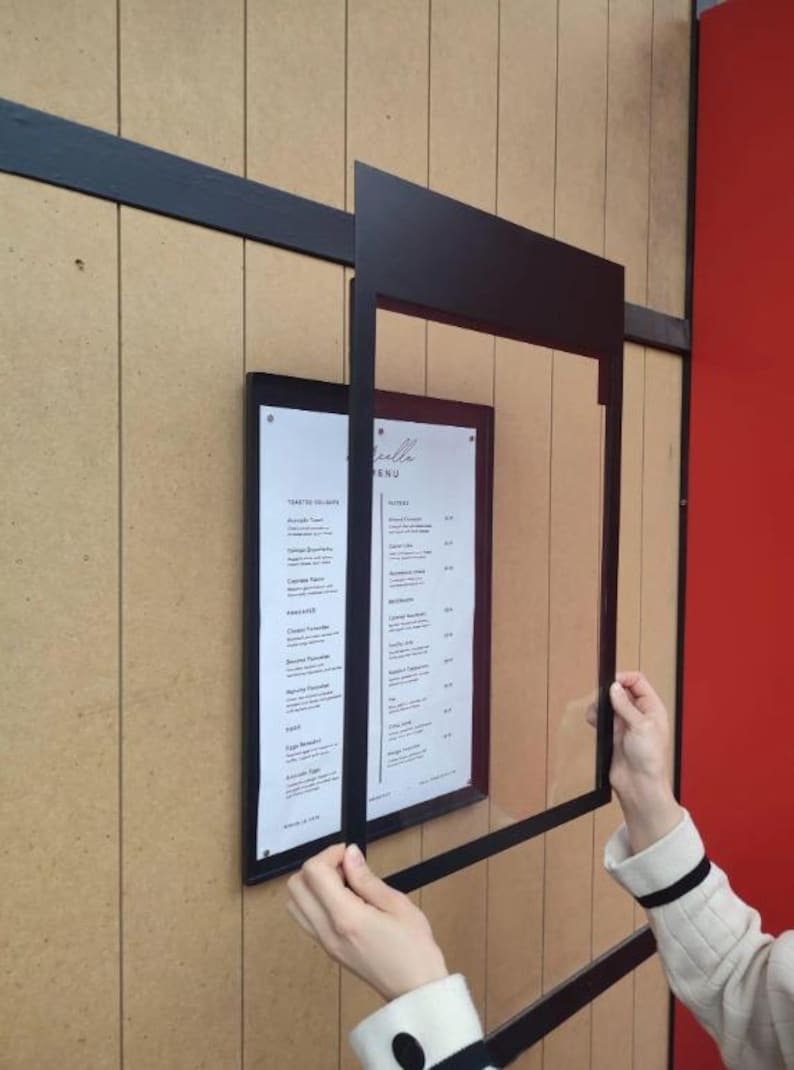 Black wall mounted menu frame with a removable clear acrylic front panel being positioned over a printed menu. Practical restaurant menu display case designed for updating food selections, seasonal menus and price lists.